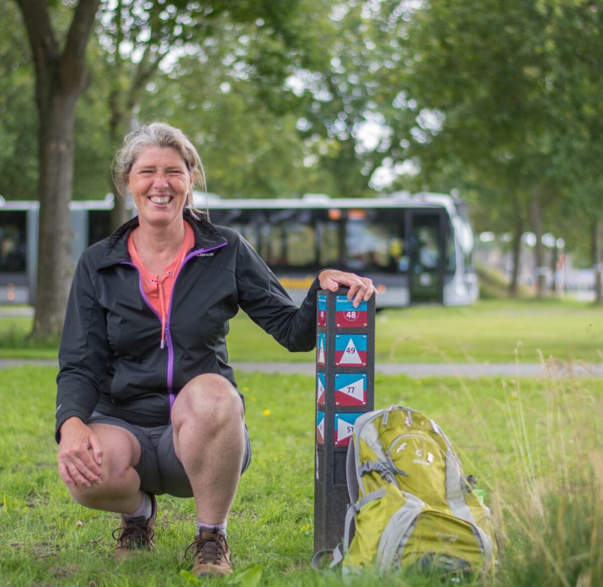 Detty maakt als fitstap-coach in opleiding gebruik van de wandelroutes via hubs. Zij zit geknield bij een routepaaltje van Wandelnetwerk Groningen. Achter haar rijdt een bus.